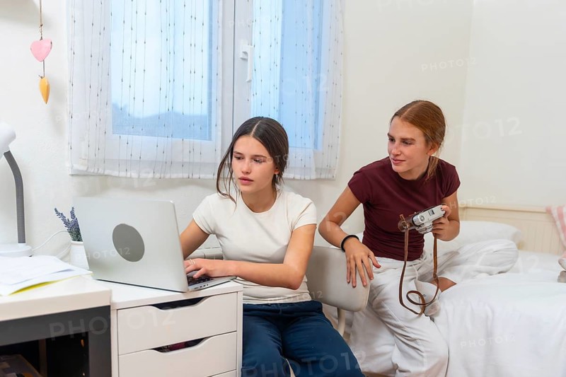 Teenage sisters sharing a bedroom, one focusing on studying or browsing online using a laptop while the other is holding a camera, ready to engage in her photography hobby