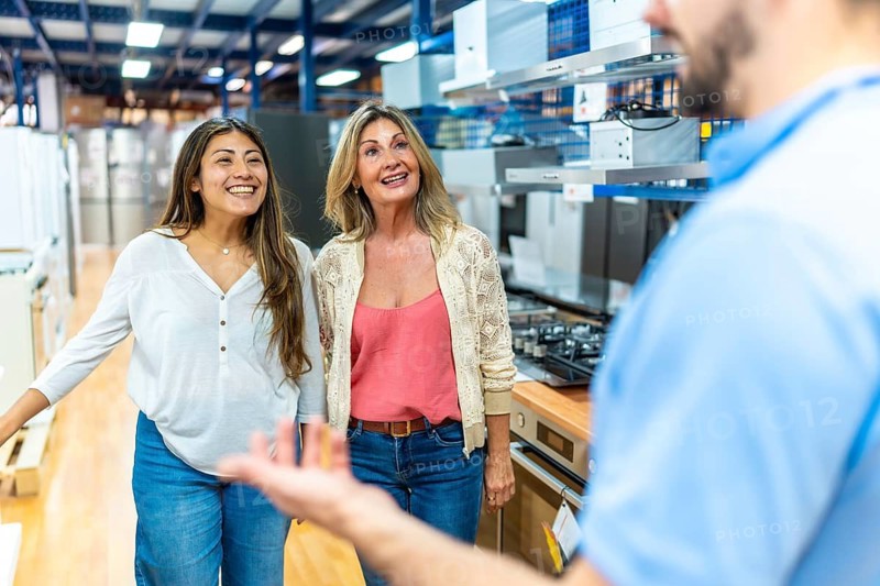 Mother and adult daughter looking at kitchen appliances and listening to a salesperson in a home improvement store, considering options for their renovation project