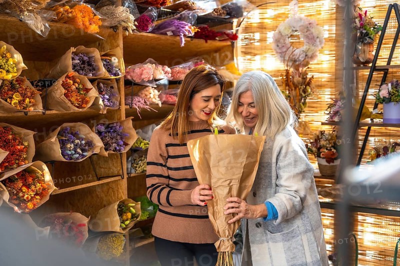 Smiling mother and daughter in a cozy flower shop sharing joy while arranging and selling artisanal dried bouquets, showcasing small business teamwork and creative connection
