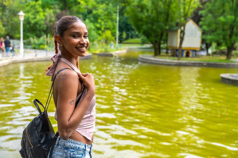 Young woman with a backpack smiling, standing by a calm green pond in an urban park, enjoying a day outdoors with a feeling of relaxation and happiness