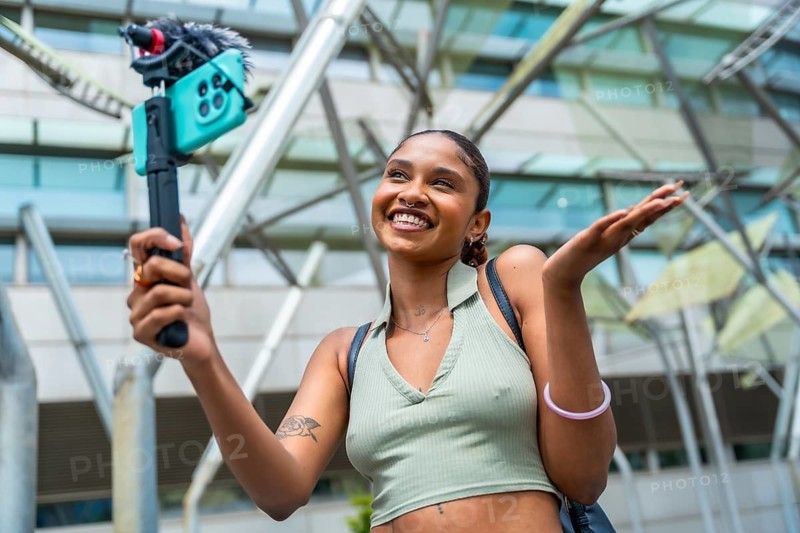 Young content creator recording herself with a smartphone on a gimbal, smiling and gesturing while vlogging in an urban environment, sharing travel and lifestyle experiences