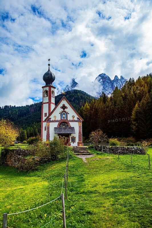 Picturesque church against an autumn mountain backdrop, surrounded by forests and green meadows under cloudy skies, Church of St. Johann in Ranui, Villnöss Valley, South Tyrol, Italy, A small church in the Alps surrounded by autumnal landscape and dramatic cloudy skies, Church of St. Johann in Ranui with Geisler Group, Villnöss Valley, South Tyrol, Italy
