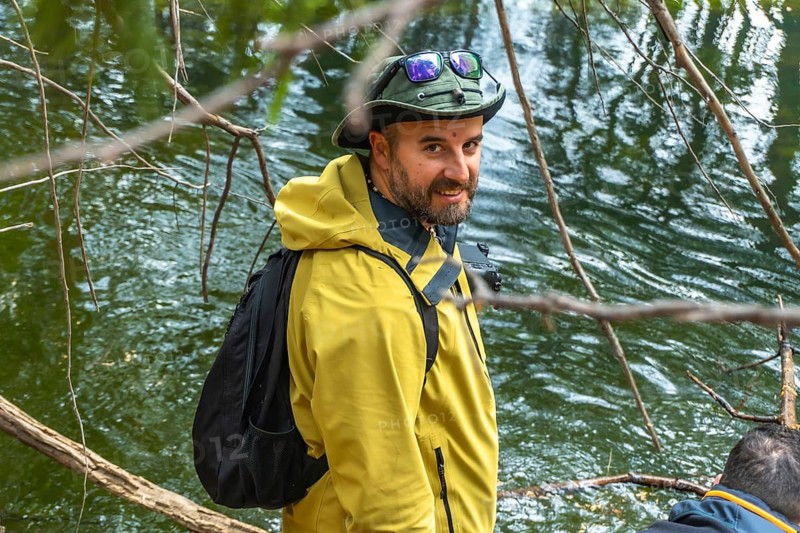 Man in outdoor clothing and hat with sunglasses worn on top, carrying a backpack and looking at the camera while standing beside a river in a natural forest environment