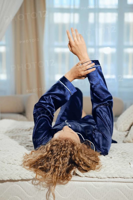 Woman lies on soft bed with arms lifted above head. Bright light enters through large windows. Hair is curly and loose. Room has light-colored curtains and bedding