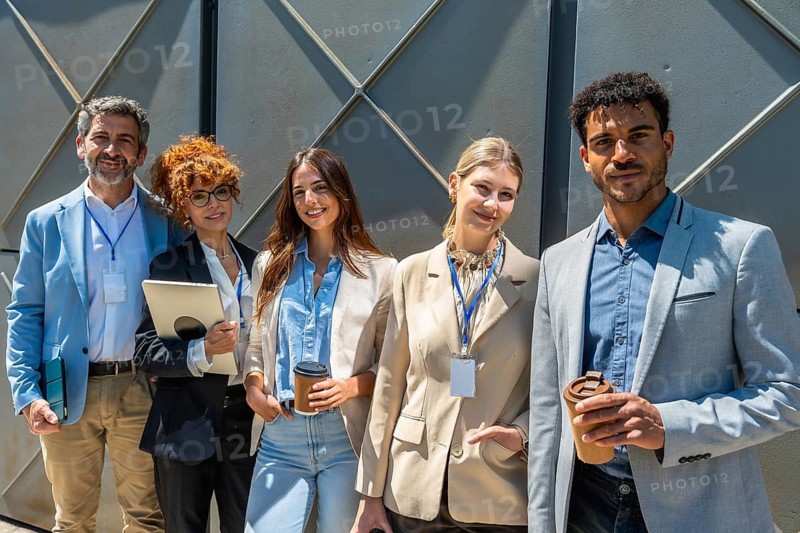 Group of five adult professionals standing outdoors, smiling towards the camera, representing collaboration, diversity, and successful team work in a corporate environment