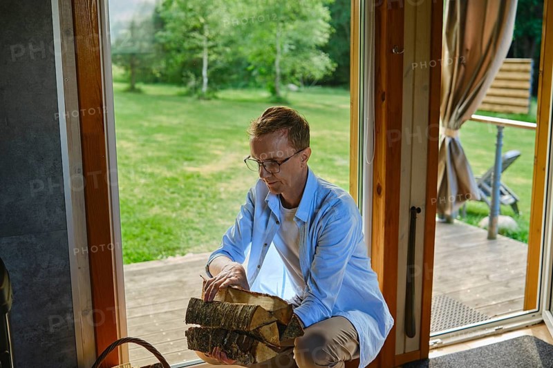 Middle-aged man stacks firewood near stove in wooden cabin. Green trees and lawn are visible in background