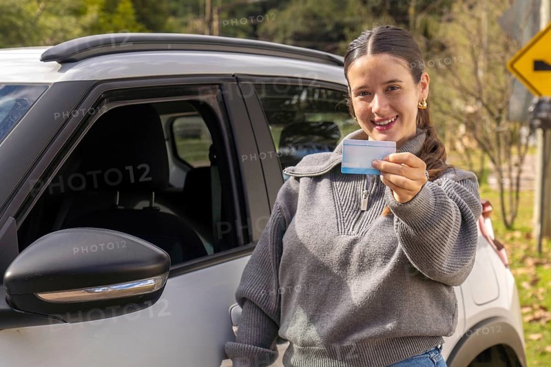 Young woman standing beside white car holding driving license, road curve warning sign visible in background, rural road setting