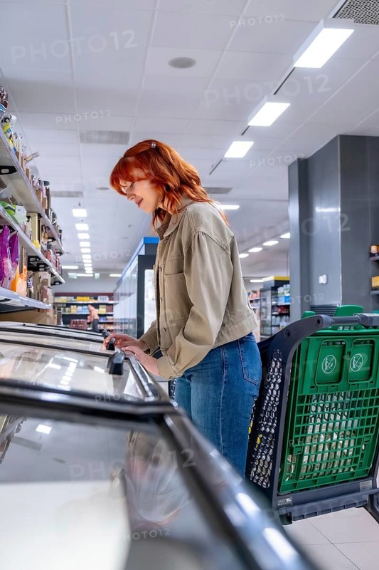 Redhead woman pushing a shopping cart and selecting items from a freezer display in a brightly lit grocery store, focusing on purchasing frozen products