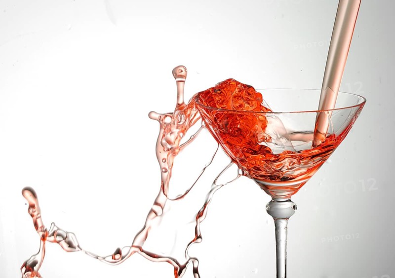 A powerful flood of red liquid pours into a cocktail glass and spills over, studio shot against a white background