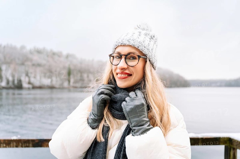 Smiling woman outdoors on a cold winter day near a snowy forest and lake. A woman wearing a white winter coat, knitted hat, scarf, gloves and glasses stands outdoors near a snowy lake, adjusting her scarf with a cheerful, relaxed expression. Snowflakes rest on her clothing, creating a bright winter moment that highlights seasonal fashion, outdoor lifestyle and cold-weather travel
