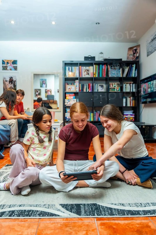 Three smiling young girls sitting on the floor, using a digital tablet together, sharing a moment of connection and entertainment in a cozy home environment