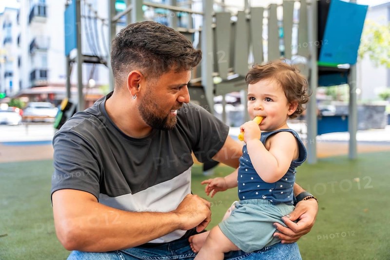 Father holding his infant son in a playground, the happy toddler enjoying a healthy snack during family time outdoors, focusing on carefree childhood and parental care