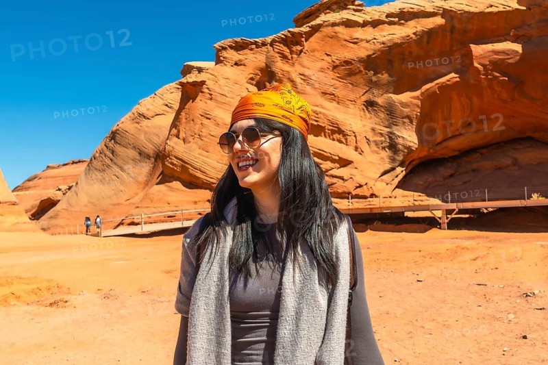 Young woman smiling and wearing sunglasses with an orange bandana on her head, enjoying a desert adventure at biidi the arch cave, a sandstone rock formation