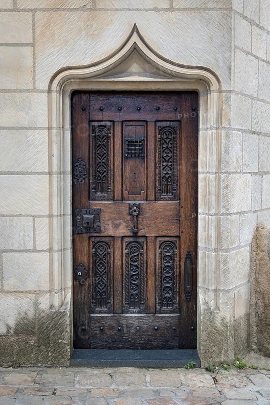 Close-up of a closed, antique entrance door made of dark natural wood with ornate decorations in the old town of Angers. The entrance is surrounded by a bright, curved stone keel arch, which shows architectural details of the Gothic style. Angers, Maine-et-Loire, Pays de la Loire, France