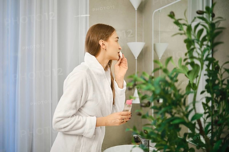 A woman stands in a bright bathroom with a mirror while applying a skincare product to her face. She holds a bottle in one hand and focuses on her reflection. A plant sits nearby on the counter