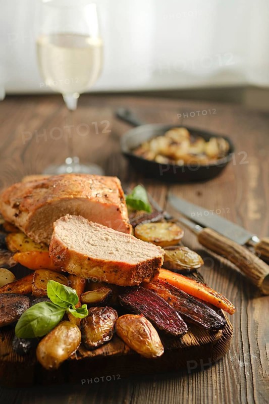 Cooked pork meat sliced and placed on a wooden board with colorful roasted vegetables. A glass of white wine is in the background. A skillet with cooked food is also visible in the setting
