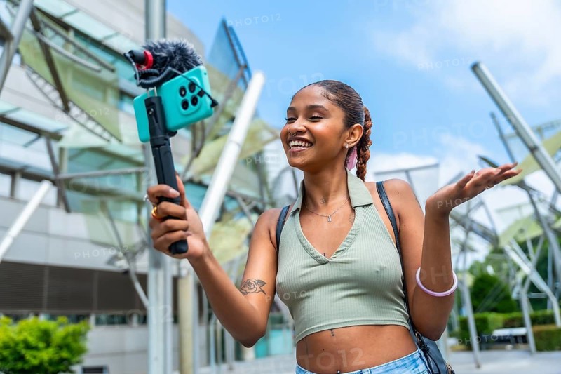 Young content creator recording video for her channel, actively engaging with her audience using a smartphone mounted on a gimbal with an external microphone in an urban setting