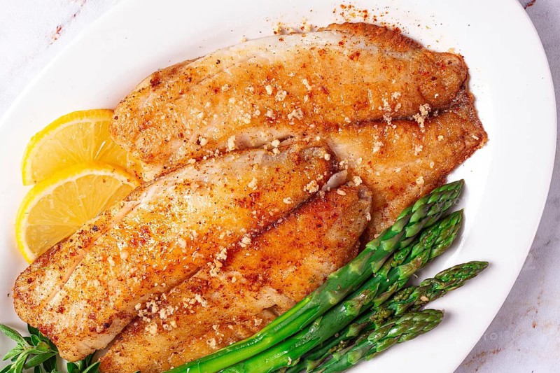 Fresh fish filets are served on a white plate alongside green asparagus and slices of lemon. The dish is prepared in a kitchen environment, ready to eat