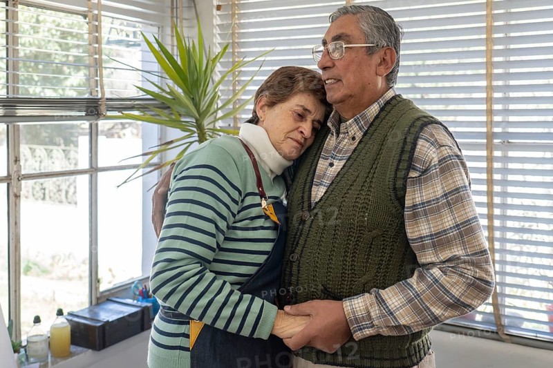 Mature woman rests her head against her partner's chest in a quiet, tender embrace inside their home art studio, holding hands gently