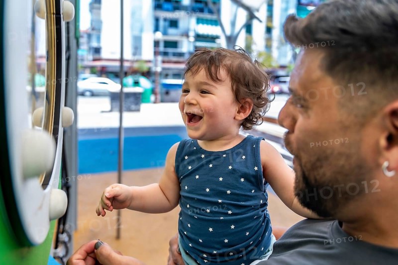 Toddler son laughing in father's arms during playful bonding at an urban park playground, candid summer moment of joy, connection and carefree family fun outdoors
