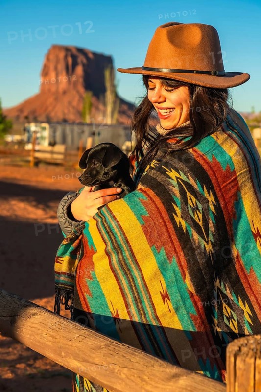 Smiling woman wearing a hat and a colorful poncho, holding a small black puppy, enjoying the desert sunrise with impressive geological formations in the background