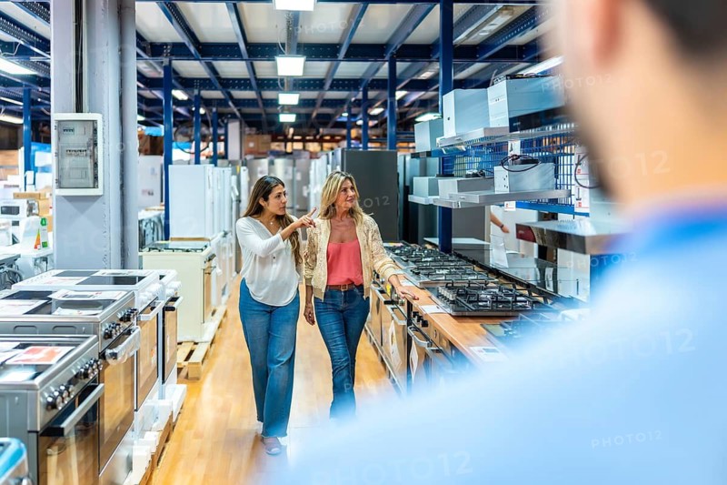 Two women walking through an aisle of a home improvement store, looking at kitchen appliances and choosing products for their home, with other customers visible in the background