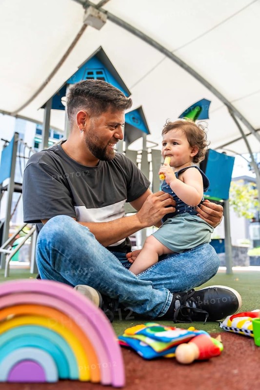 Happy father smiling and interacting with his young son on a playground, with colorful toys scattered around them, enjoying a sweet moment of family connection and parenthood
