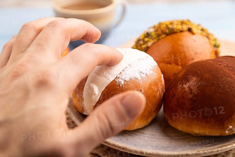 Maritozzo traditional Italian bun dessert with hand, cup of coffee, on blue wooden background with orange linen textile, close up, side view, selective focus