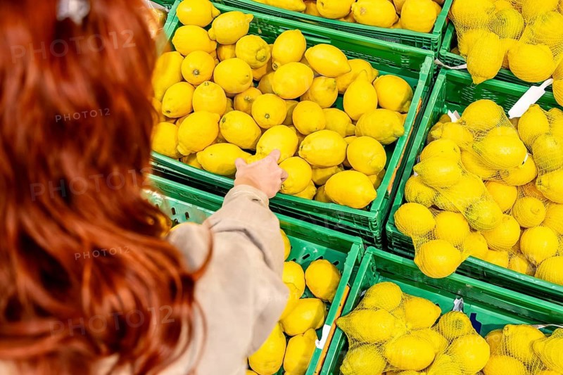 Redheaded woman selects bright yellow lemons from green crates at a supermarket produce display, close up on healthy choices and fresh citrus for cooking, drinks, wellness