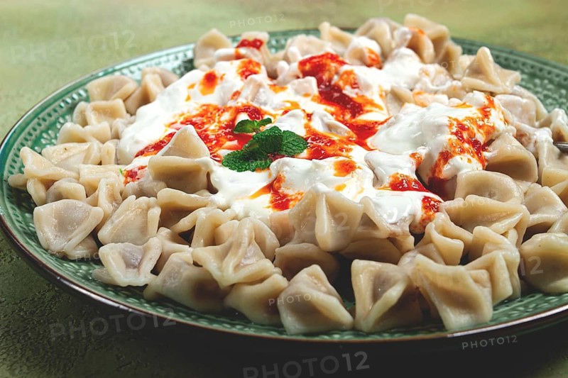 The dish features a generous serving of Turkish dumplings topped with velvety yogurt and a drizzle of spicy red sauce, displayed on a beautifully crafted plate with a green accent