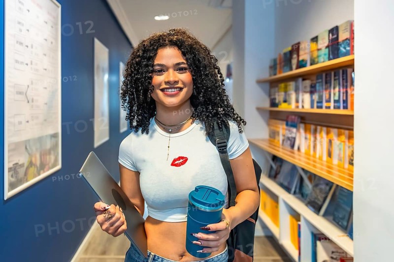 Young female student smiling at camera, holding a laptop and a reusable cup while standing in a university hallway or college library, representing education and modern learning