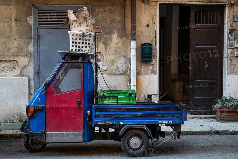 Blue Piaggio Ape, Italian three-wheeler utility vehicle, with red door standing loaded in front of a door in an old town street, Palermo, Sicily, Italy