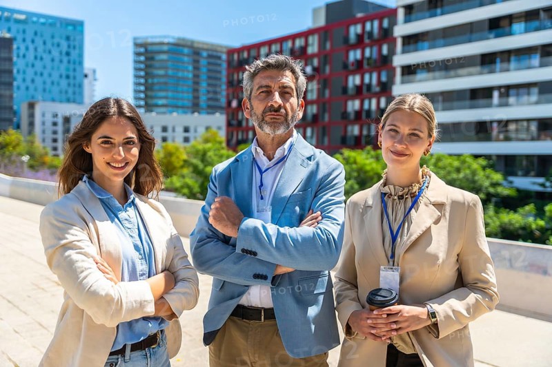 Business professionals, two women and one man, wearing smart casual attire and lanyards, standing outdoors in an urban setting with modern architecture behind them