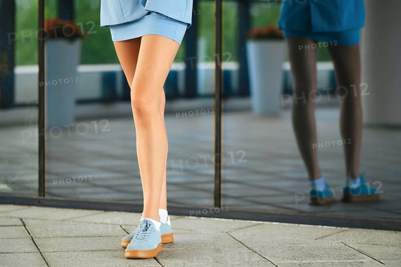 Woman with slender legs wearing blue sneakers and skirt standing close to glass wall with reflection in urban area. Flowers in pots are visible in background. The setting shows signs of a busy city