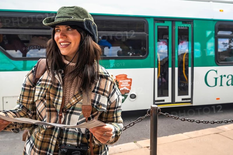 Smiling woman traveler in a plaid shirt and hat holds a park map at a shuttle bus stop, ready to explore grand canyon national park and hike scenic viewpoints