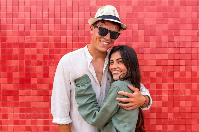 Cheerful young couple smiling and embracing each other, standing against a vibrant red tiled wall, expressing happiness and togetherness in a summer setting