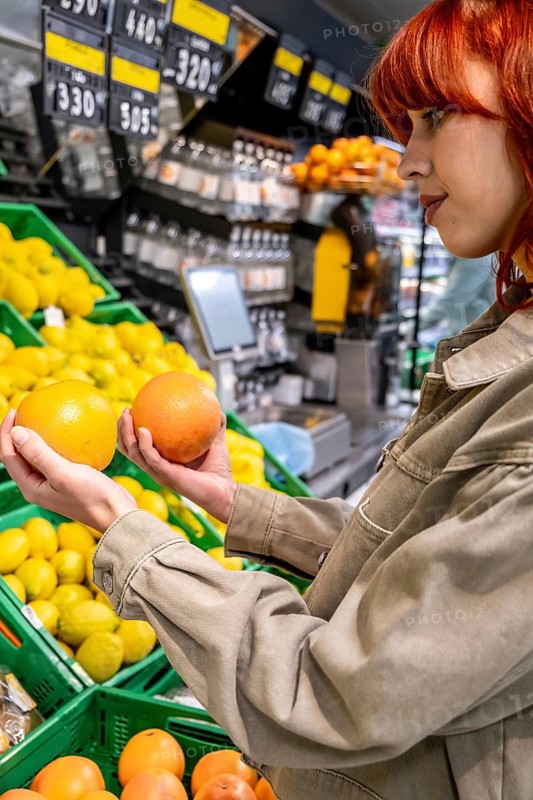 Woman carefully selecting ripe oranges and grapefruit from a large display of produce in a modern grocery store, focusing on healthy eating and shopping choices