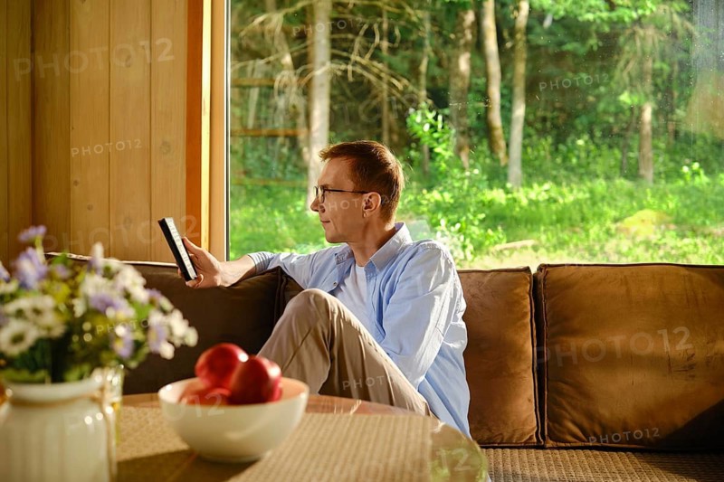 Retired man sits on couch in living room in wooden cabin holding electronic boor in hand and reading. Calm and leisurely retirement life away from noisy city and bustle