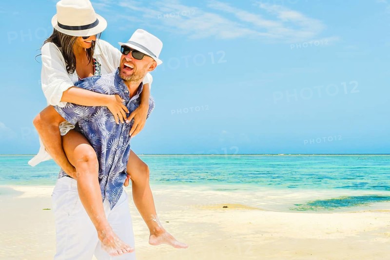 Multi-ethnic happy young couple having fun piggyback riding on white sand beach. Caucasian man and middle aged woman in playful moment on the shore at summer time - Concept of happiness and travel