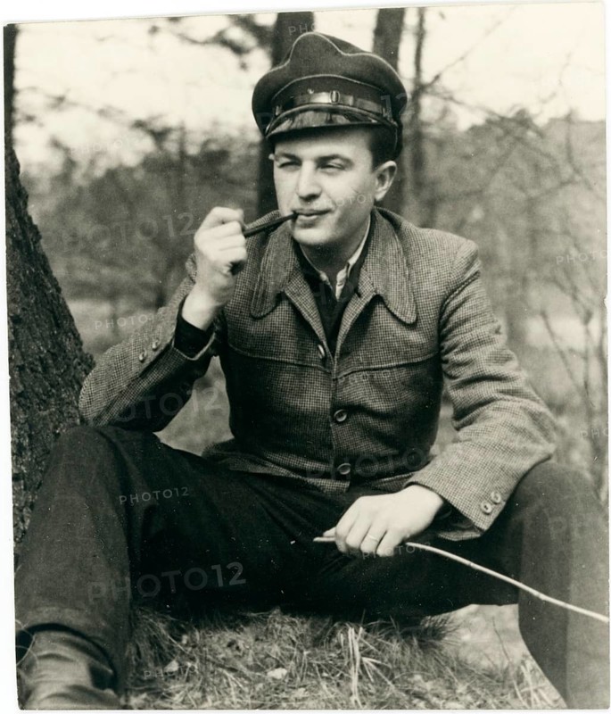 A man in uniform sits smiling with a pipe in a meadow under a tree, Herbert Jacobi (1915–1978), Self-portrait, c. 1948