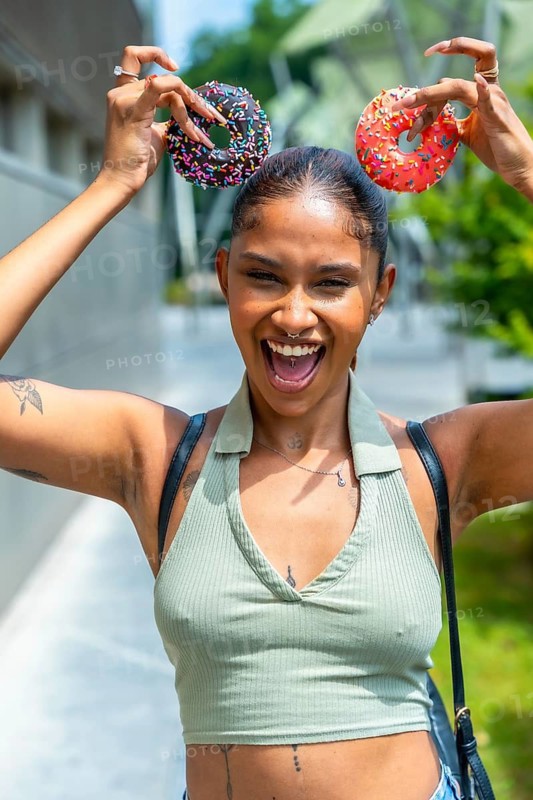 Smiling young woman making a funny face, holding two colorful sprinkle donuts like ears over her head, expressing joy and playful happiness while walking outdoors on a sunny day