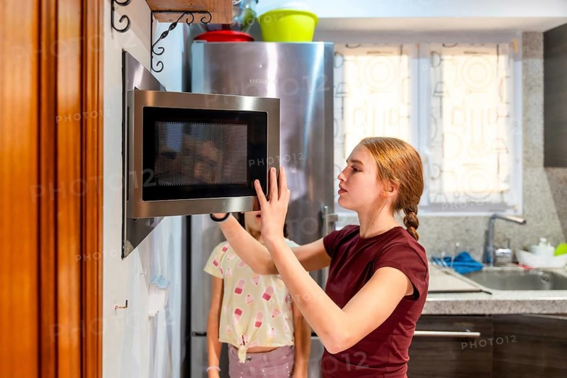 Two sisters warming food in a bright modern kitchen, sharing a casual moment while using the microwave, portraying everyday family routine and domestic life with natural warmth