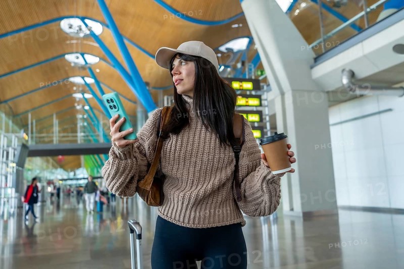 Young woman checking her smartphone and holding a coffee cup while walking through a contemporary airport, managing her journey with luggage and backpack in a busy travel environment