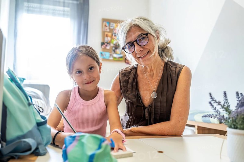 Grandmother and granddaughter are sharing a bonding moment, sitting at a desk and smiling for the camera while engaging in learning activities and studying at home