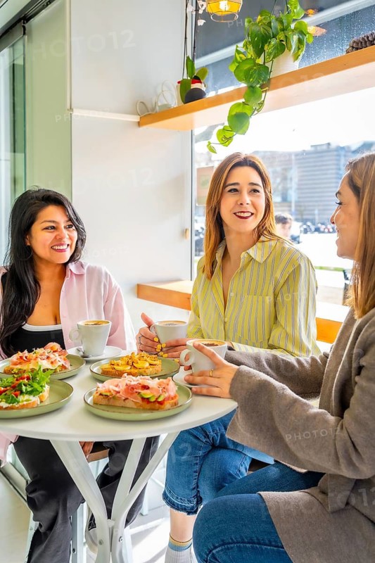 Three smiling women sitting at a table in a bright cafe, enjoying coffee and toast with different toppings, sharing a pleasant conversation and friendship over brunch