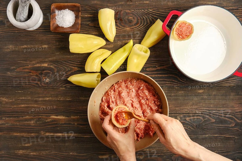 Hands are stuffing bell peppers with ground meat Nearby are halved peppers ready to be filled. Kitchen table shows some salt and cast-iron pot. Scene suggests cooking in a home setting
