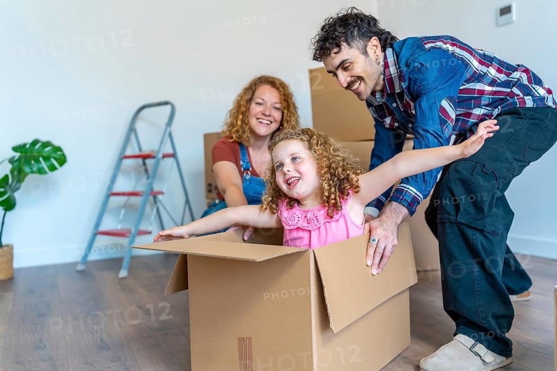 Happy family moving to a new house, father pushing cheerful daughter in a cardboard box, parents and child actively playing together while packing or unpacking belongings in a bright, modern room