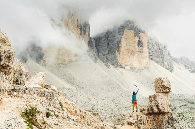 Female hiker raising trekking poles beneath misty Dolomites cliffs celebrating achievement adventure freedom and mountain exploration