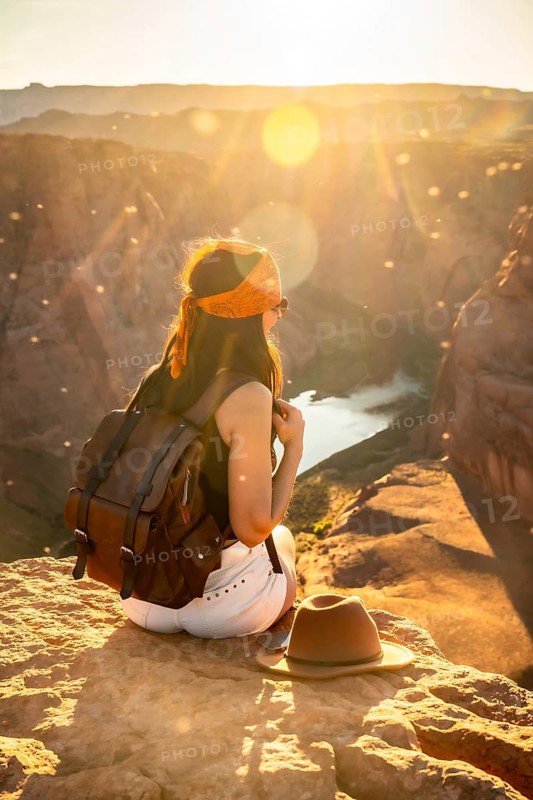 Woman traveler sitting with a backpack on a cliff edge, watching the panoramic sunset view over horseshoe bend in page, arizona, with a river winding through the desert canyon below