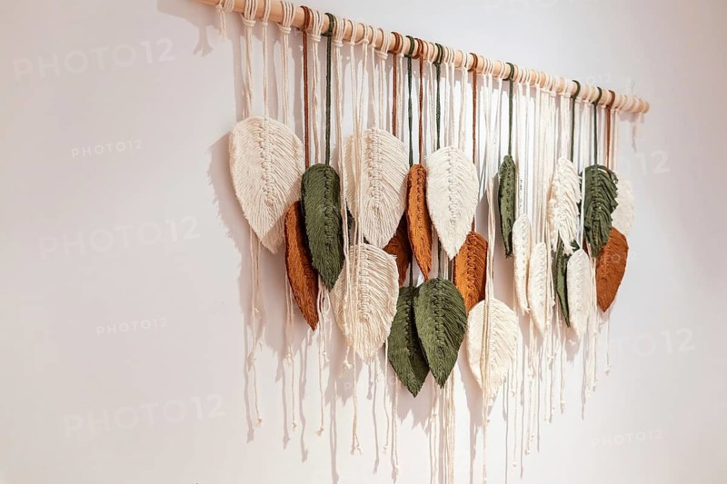 Handmade macrame wall hanging featuring cotton leaves in white, green, and brown, creating decorative bohemian fiber art for modern home decor on a simple white background